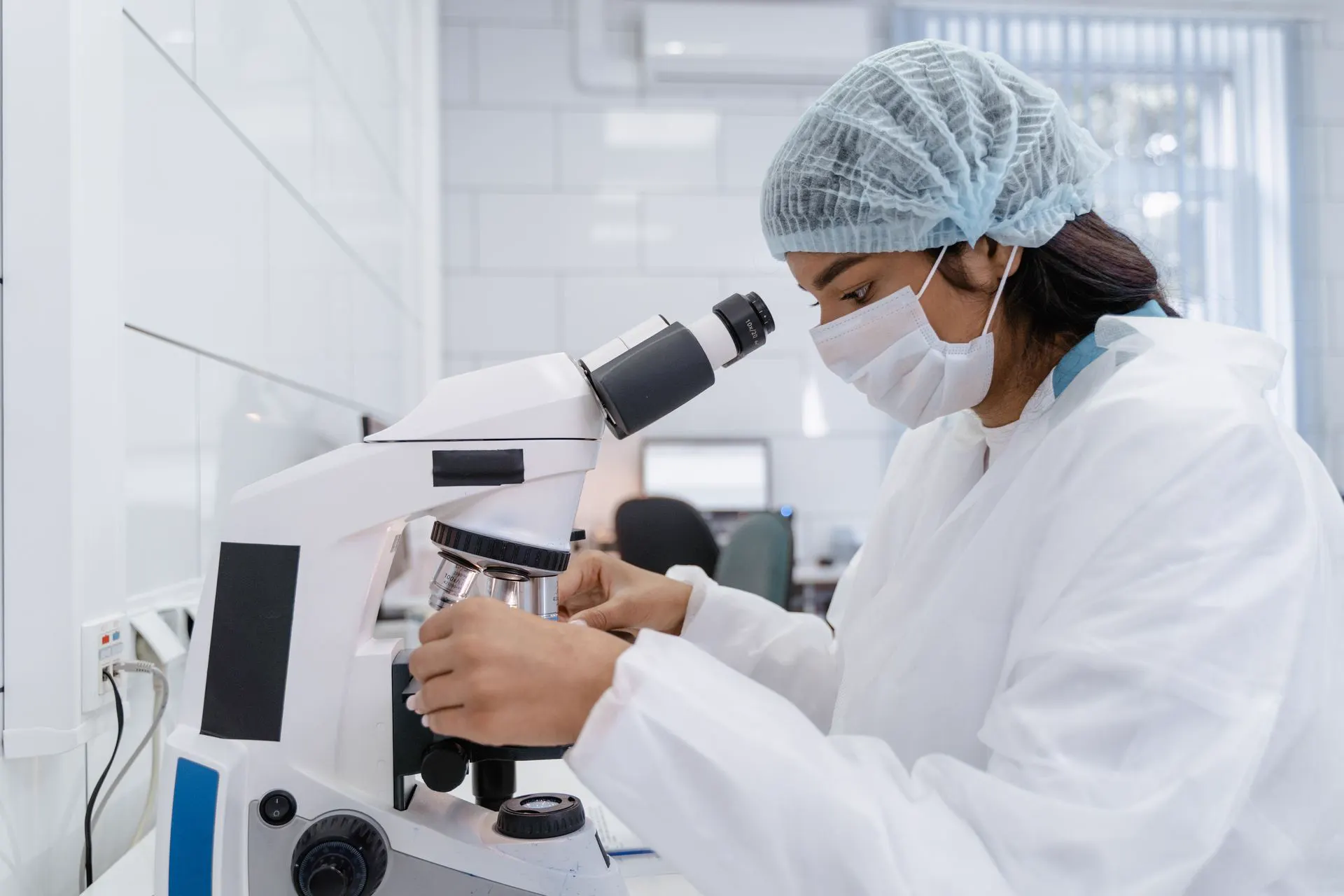 Scientist in a hairnet and mask examining a sample under a microscope